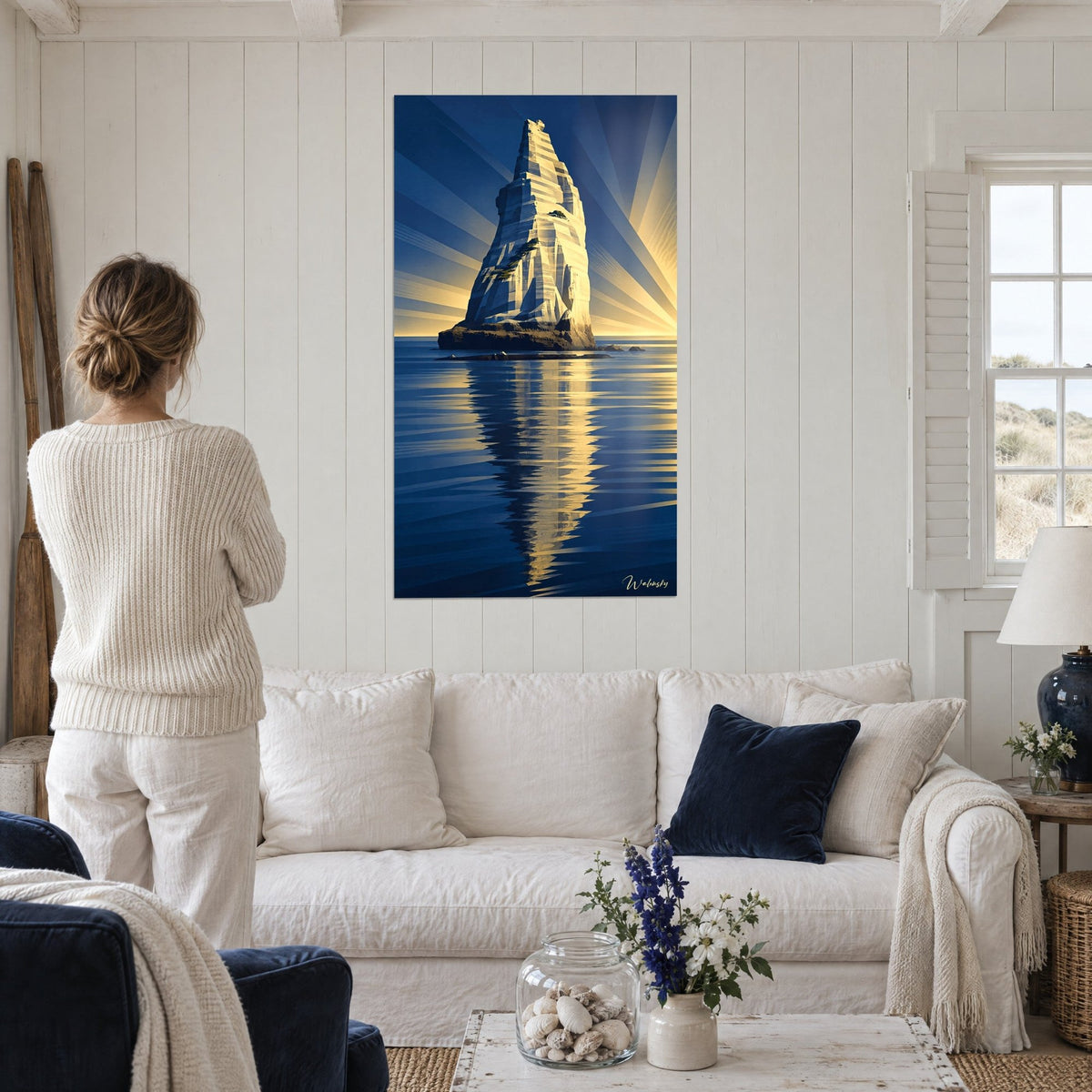 Wall art chalk needle rising through golden rays above a midnight-blue sea, woman seen from behind above a sofa in a bright living room