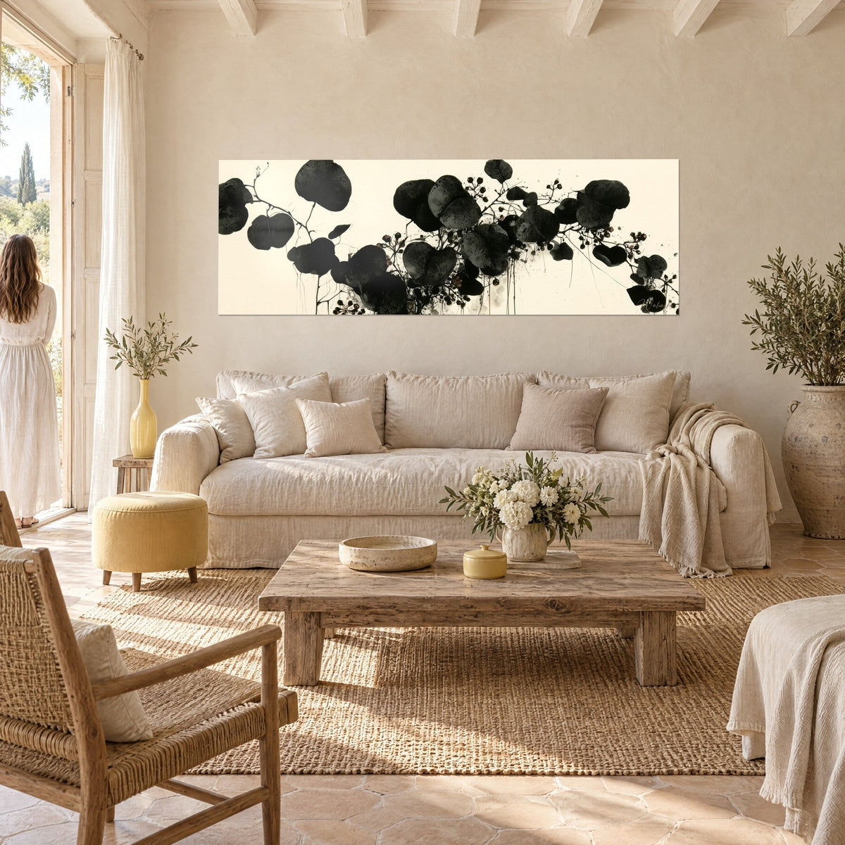 Wall art black round-leaf branch on ivory background, fine stems and berries, a woman seen from behind, above a sofa in a bright living room