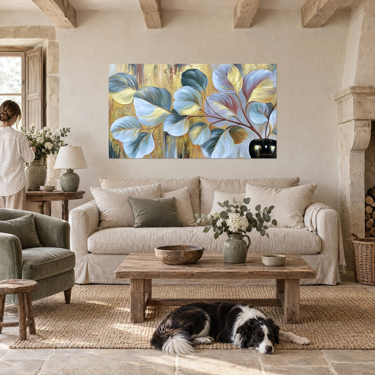 Schilderij van een eucalyptustak met zilveren en gouden bladeren, zwart gelakte vaas, liggende collie, boven een bank in een lichte woonkamer