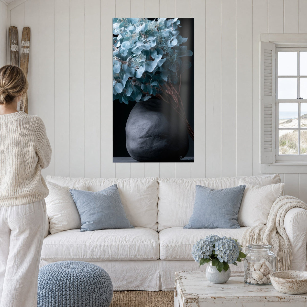 Schilderij bos lichtblauwe eucalyptus in een ronde zwarte vaas, bruine stelen, donkere achtergrond, boven een bank in een lichte woonkamer