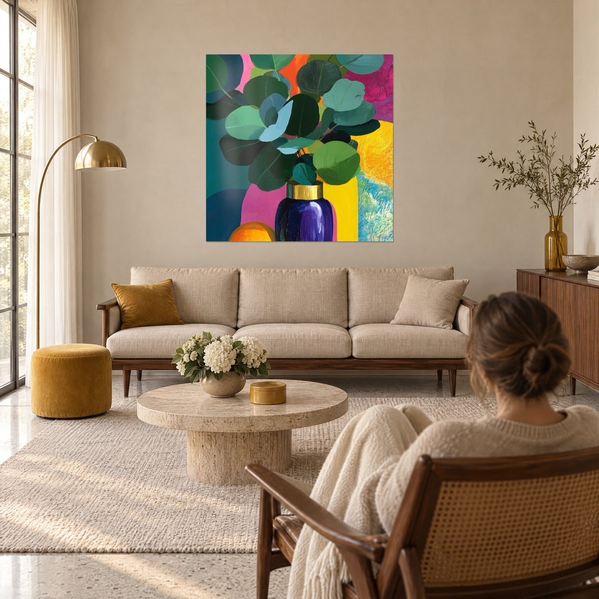 Tableau branches d'eucalyptus aux feuilles rondes vert sapin, vase bleu nuit à col doré, fond rose et jaune, au-dessus d'un canapé dans un salon lumineux