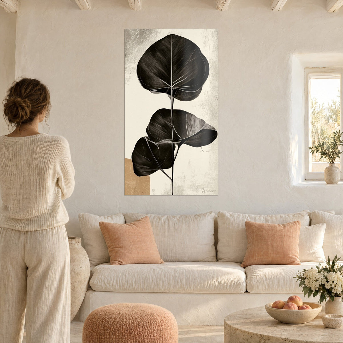 Quadro foglie nere di eucalipto dalle nervature chiare su sfondo crema e ocra, donna di spalle, sopra un divano in un soggiorno luminoso