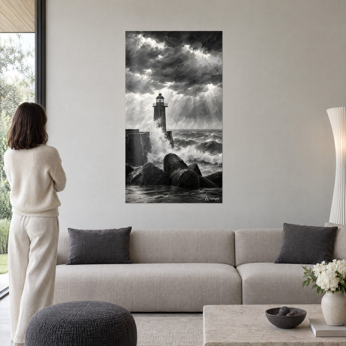 Schilderij stenen vuurtoren geteisterd door een witte golf, zwarte en grijze lucht, vrouw van achteren, boven een bank in een lichte woonkamer
