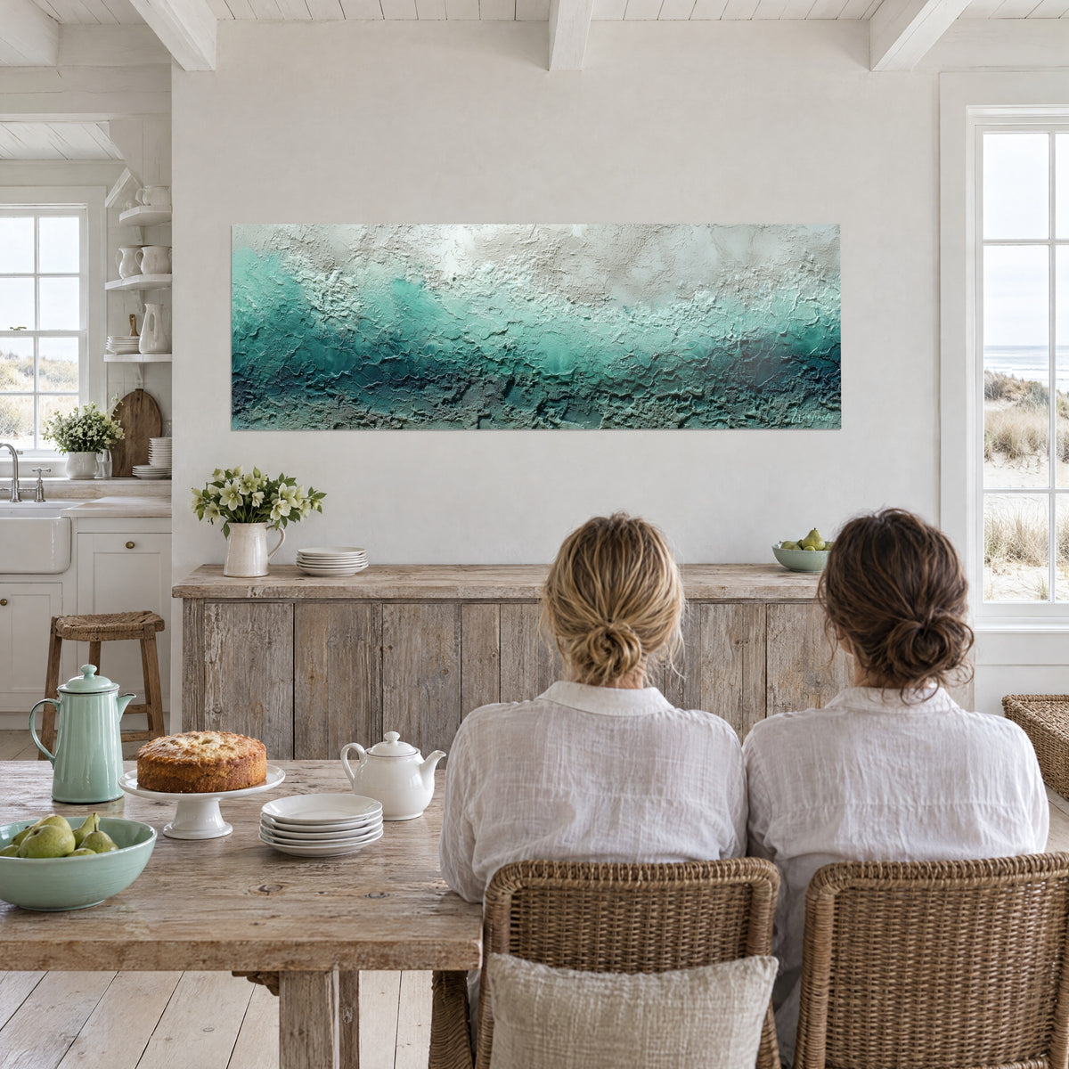 Wall art of sculpted shoreline in impasto, bright turquoise on grey-white, two women seen from behind at a table, beach house kitchen