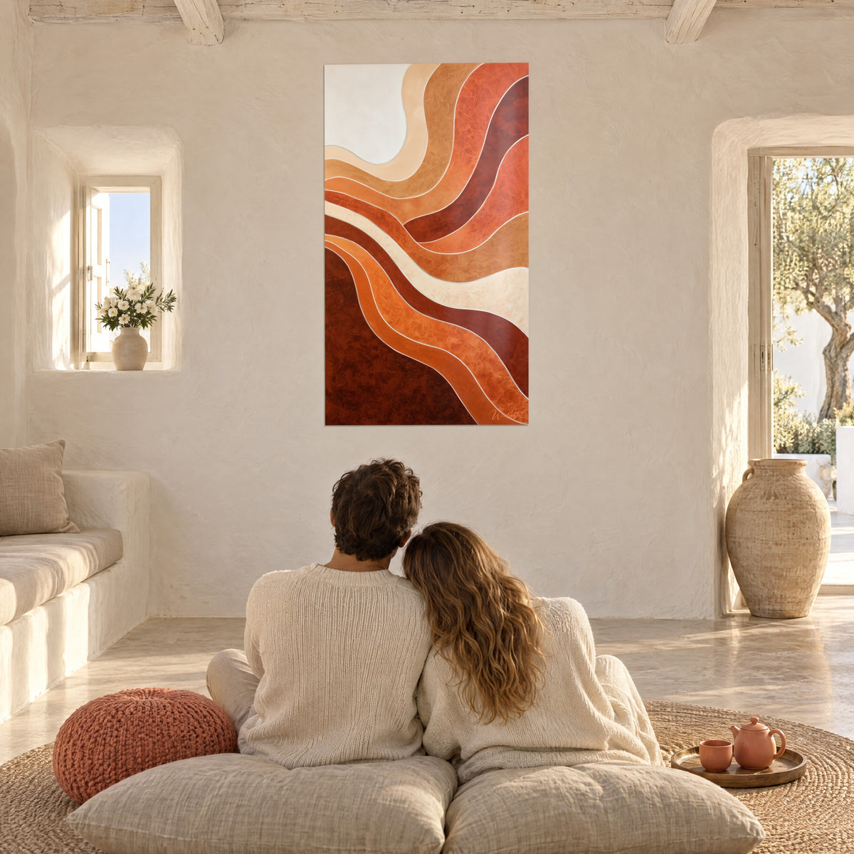 Cuadro de capas onduladas ladrillo y arena, pareja abrazada de espaldas sobre cojines, puf coral, salón mediterráneo blanco