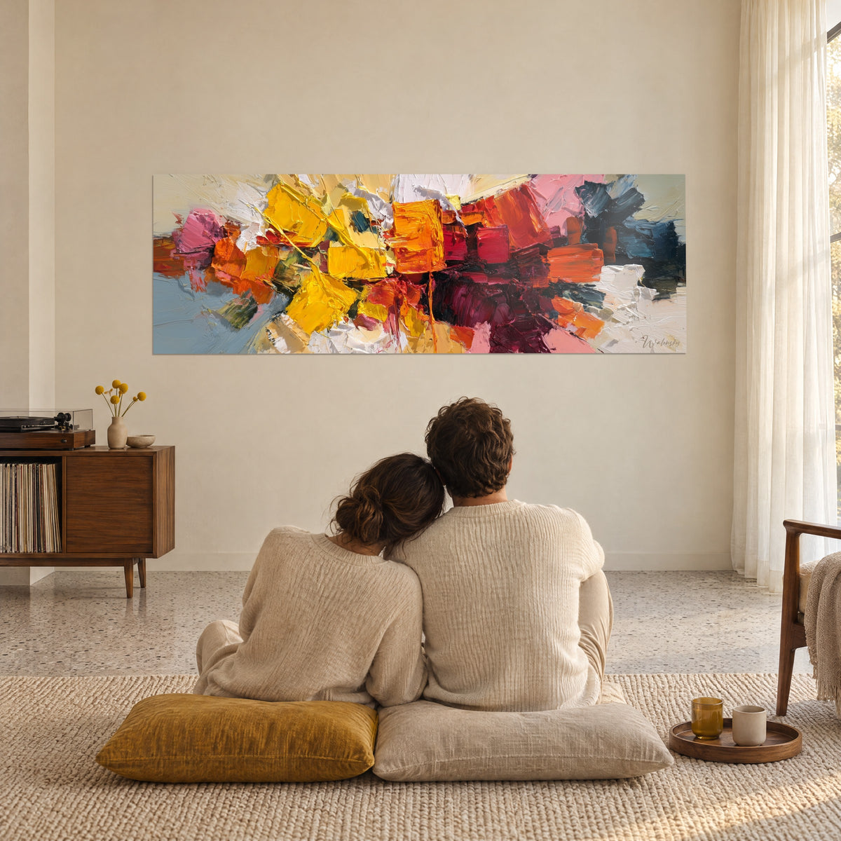 Cuadro mosaico de materia amarilla, rosa y burdeos contemplado de espaldas por una pareja sentada sobre cojines mostaza