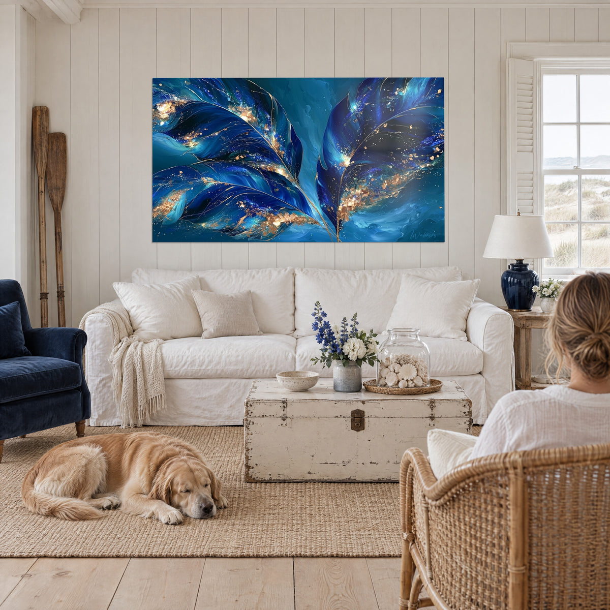 Tableau feuilles de bananier bleu marine et or aux nervures dorées, deux femmes de dos et retriever, au-dessus d'un canapé dans un salon lumineux