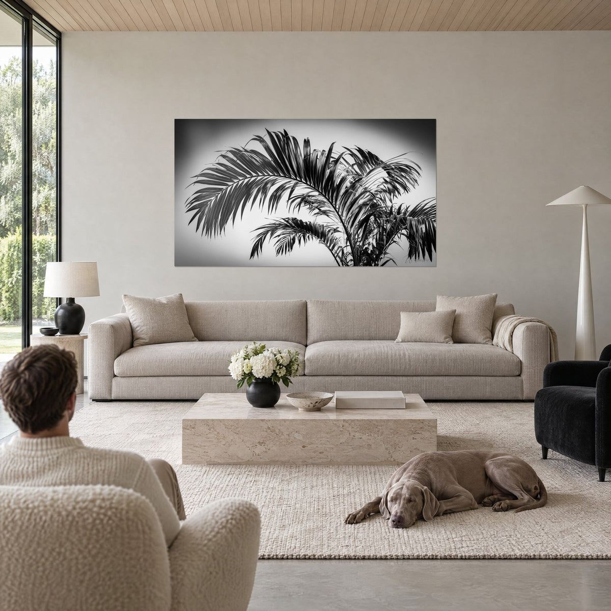 Wall art black and white palm, arched fronds with dark veins on grey background, man seen from behind and dog above a sofa in a bright living room