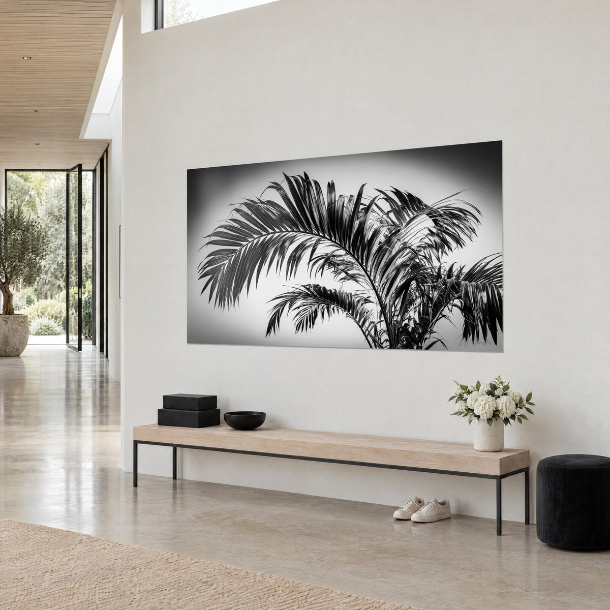 Wall art palm leaf with glossy black blades on pale grey sky, soft vignetting, white wall and light wood bench