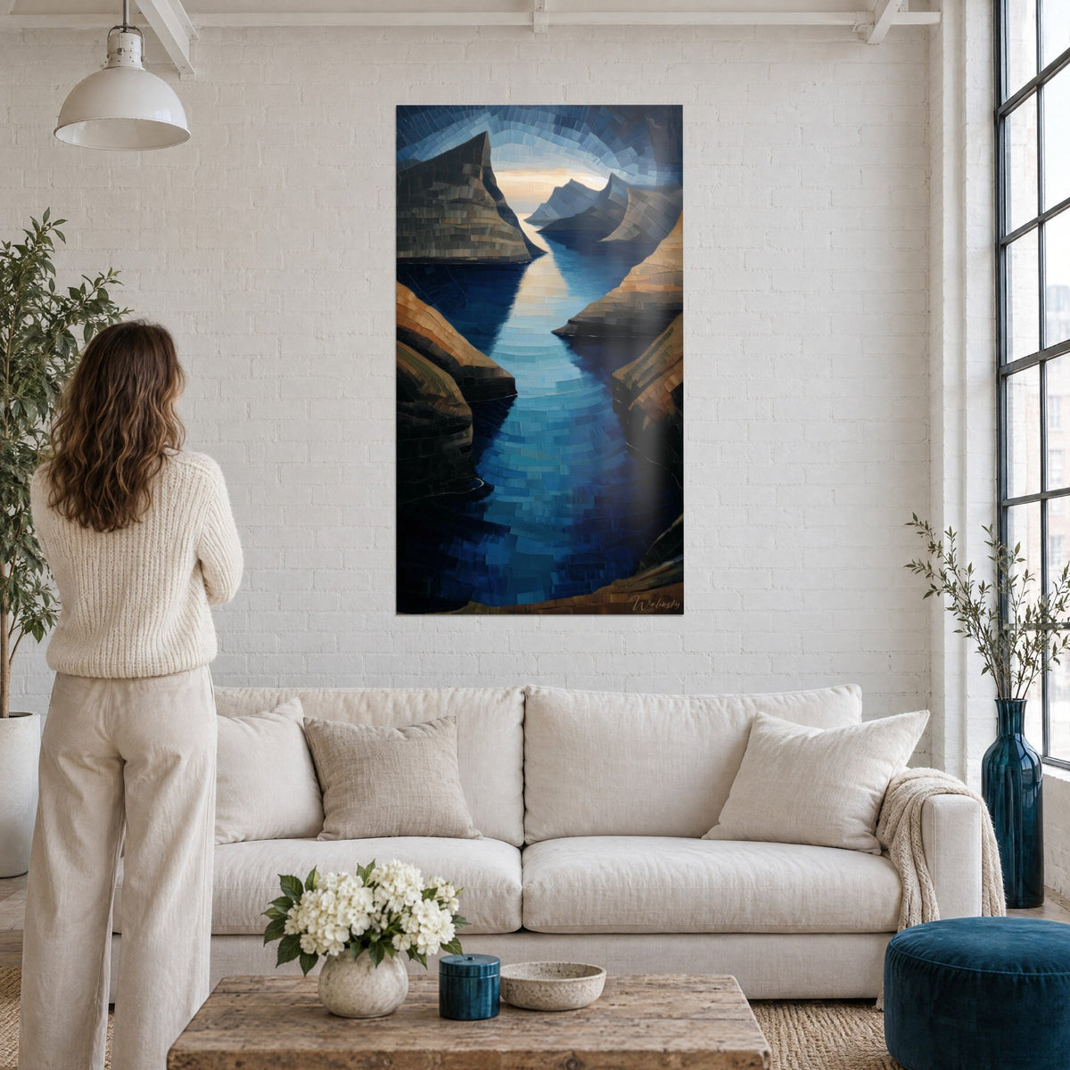 Schilderij Scandinavisch fjord met bruine kliffen en middernachtblauw water, een vrouw van achteren boven een bank in een lichte woonkamer