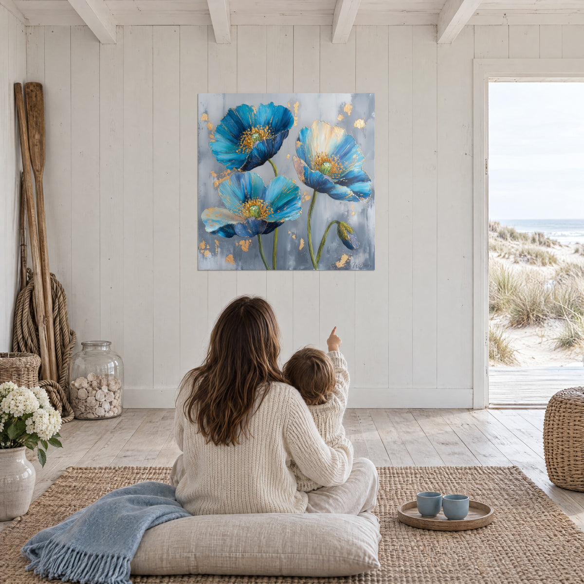 Schilderij van een klaproosbloei in blauw en goud op parelgrijs, een moeder en haar kind van achteren zittend op een kussen, huis aan zee