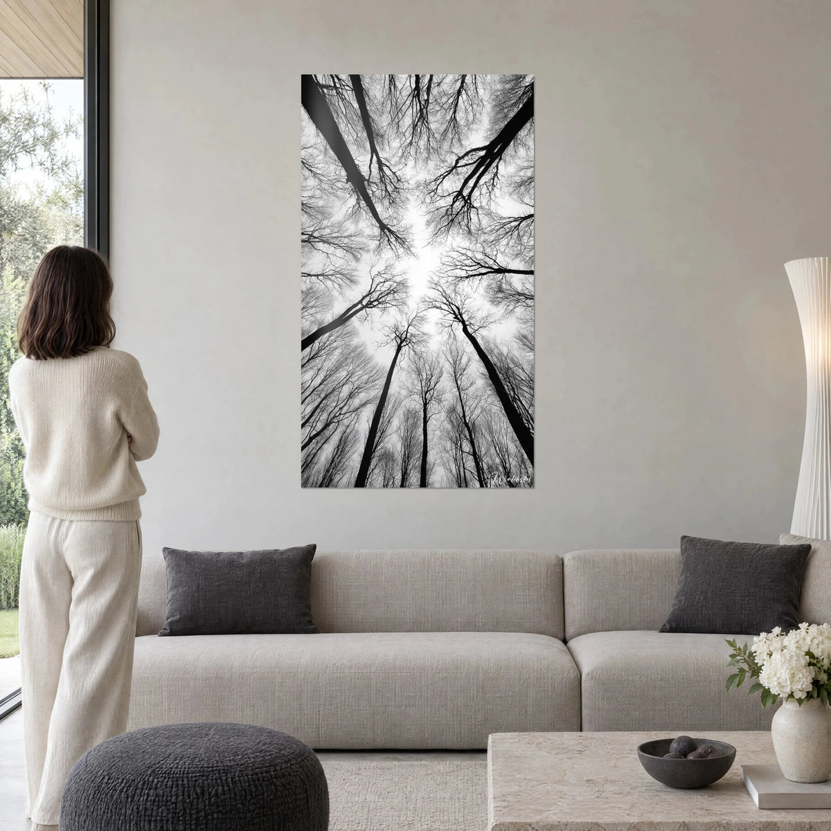 Wall art worm's-eye view of bare tree canopy, black trunks against white sky, woman seen from behind above a sofa in a bright living room