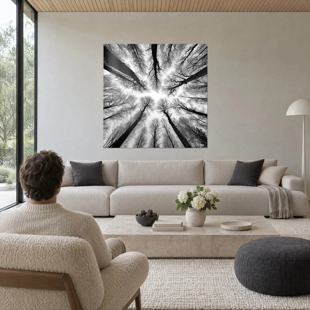 Tableau forêt vue d'en bas, troncs noirs et ciel blanc, homme de dos, au-dessus d'un canapé dans un salon lumineux