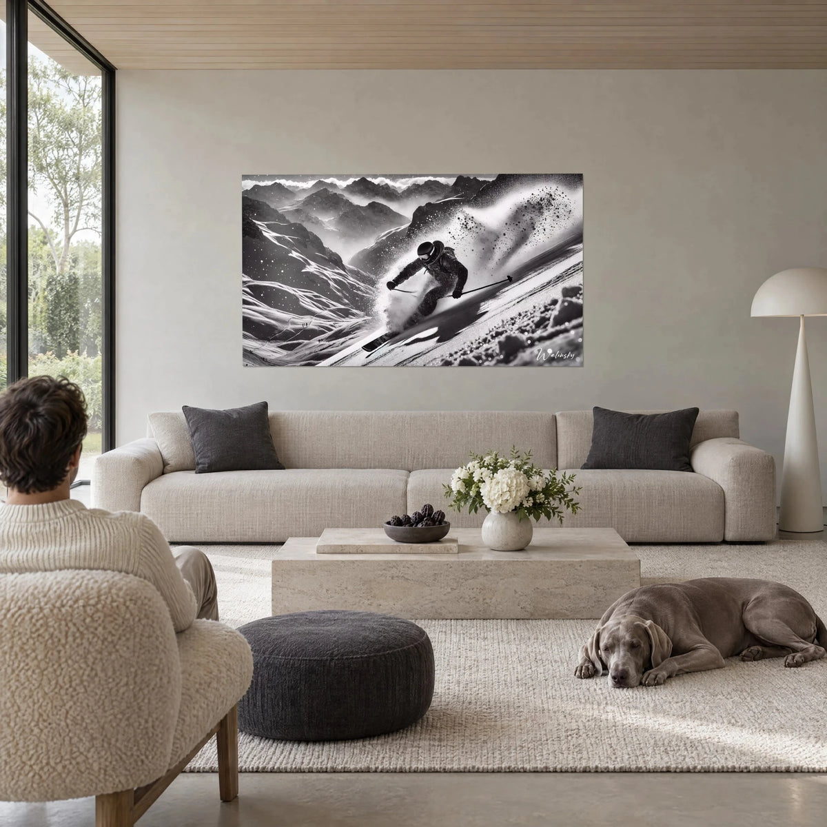 Wall art of a skier launching through powder, white spray and grey ridges, man and grey dog above a sofa in a bright living room