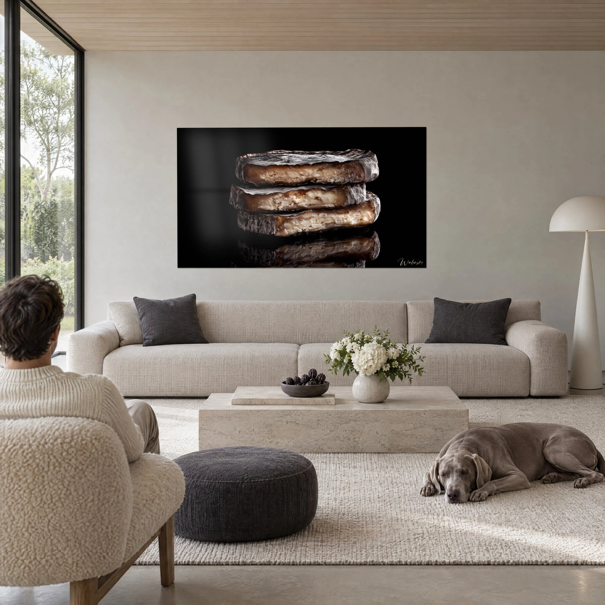 Wall art three stacked wheels of cheese with ash-grey rind and ivory paste, black background, reclining Weimaraner, above a sofa in a bright living room, Walensky