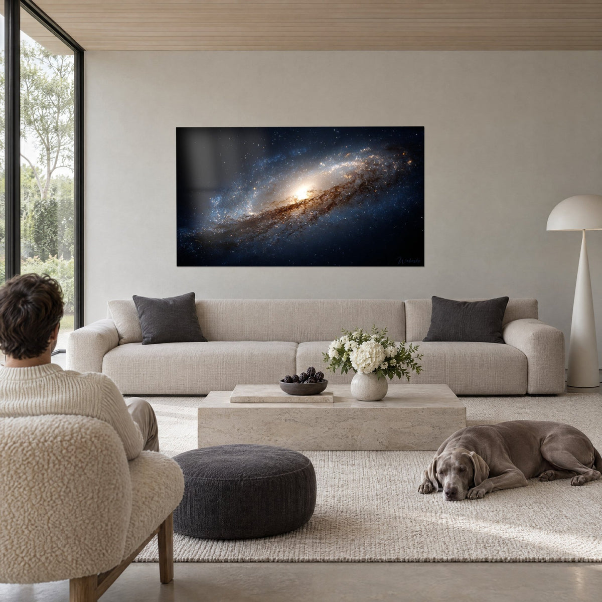 Schilderij spiraalvormige melkweg met gouden kern, blauwe armen en bruine stofwolken, twee personen met de rug naar de kijker, een Weimaraner die op een bank ligt in een lichte woonkamer
