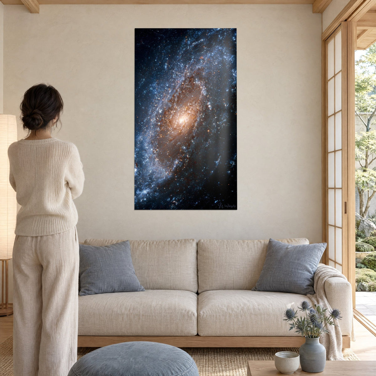 Wall art spiral galaxy with a golden core, glacier-blue arms dotted with stars, woman seen from behind, above a sofa in a bright living room