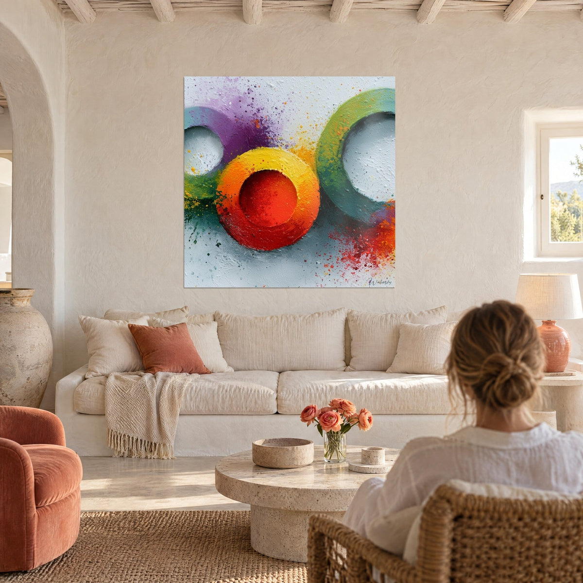 Cuadro con anillos de pintura roja, amarilla y verde salpicados, una mujer de espaldas lo contempla sobre un sofá en un salón luminoso