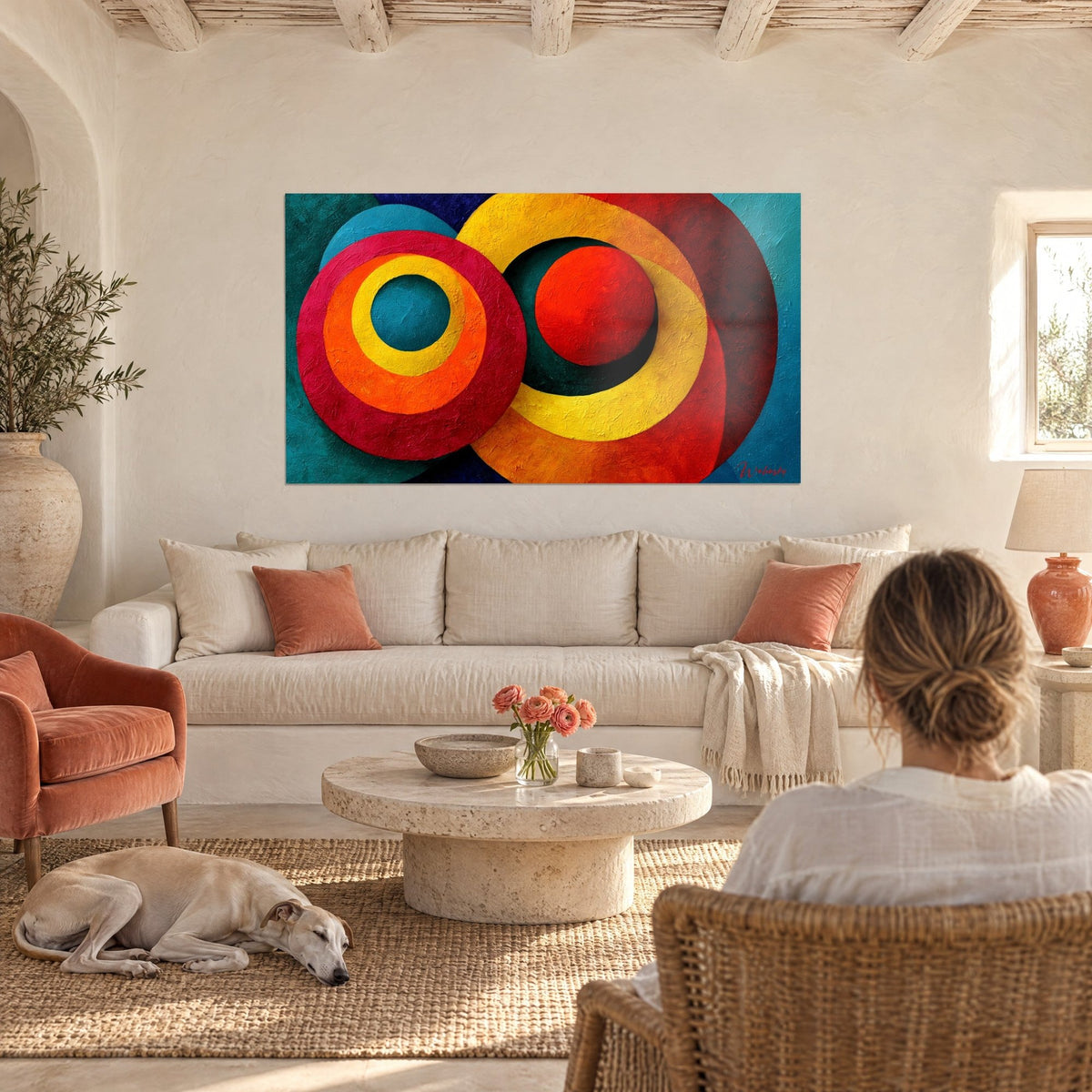 Wall art with concentric circles in carmine and lemon yellow on a teal background, a person seen from behind and a greyhound, above a sofa in a bright living room