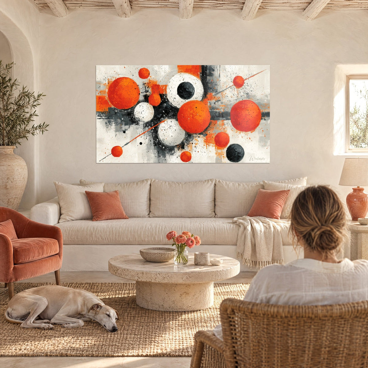 Wall art with vivid orange and white spheres splashed with black, two figures seen from behind and a sleeping greyhound, above a sofa in a bright living room