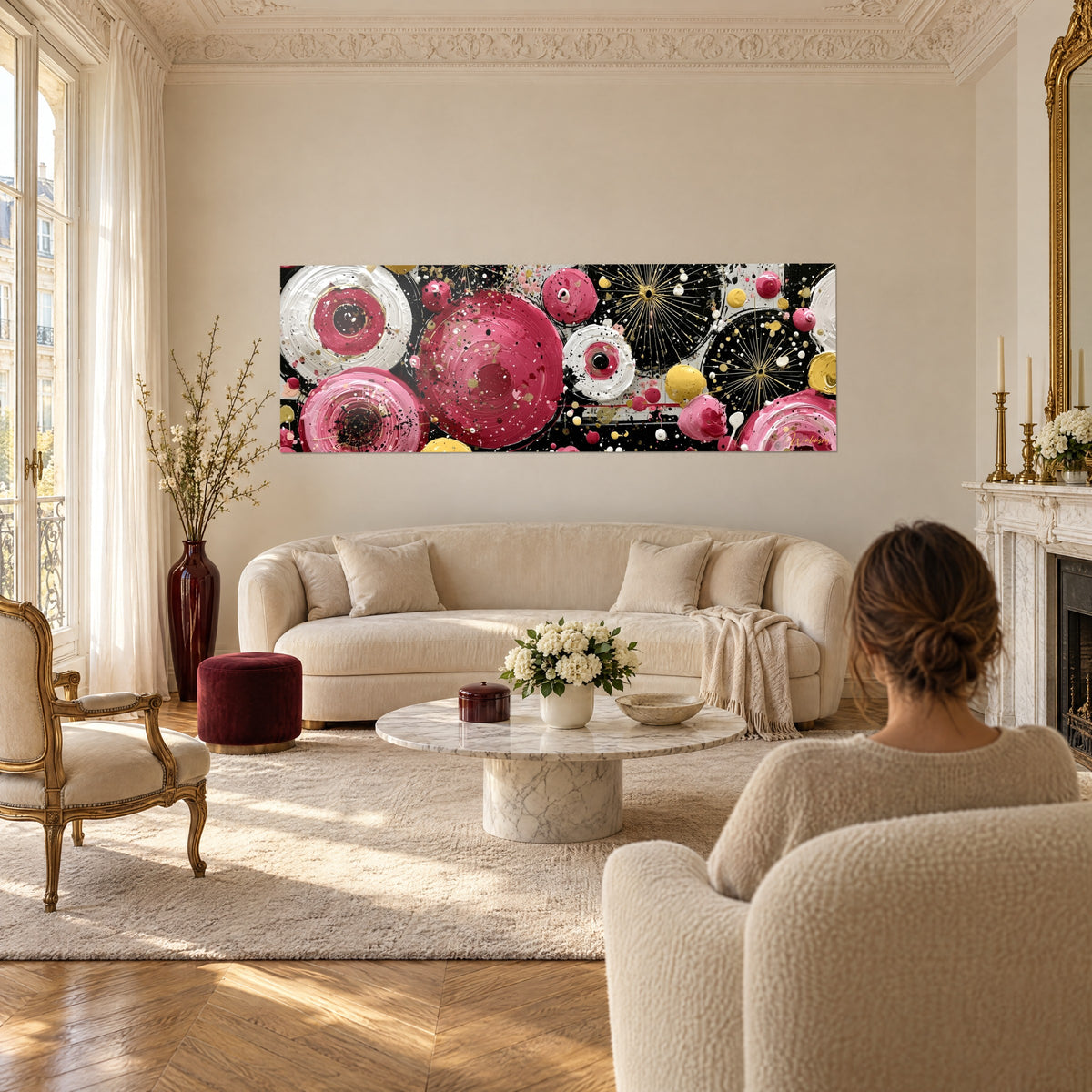 Quadro con dischi lampone e bianchi cerchiati di nero, scintille dorate, di fronte a una donna di spalle, sopra un divano in un soggiorno luminoso