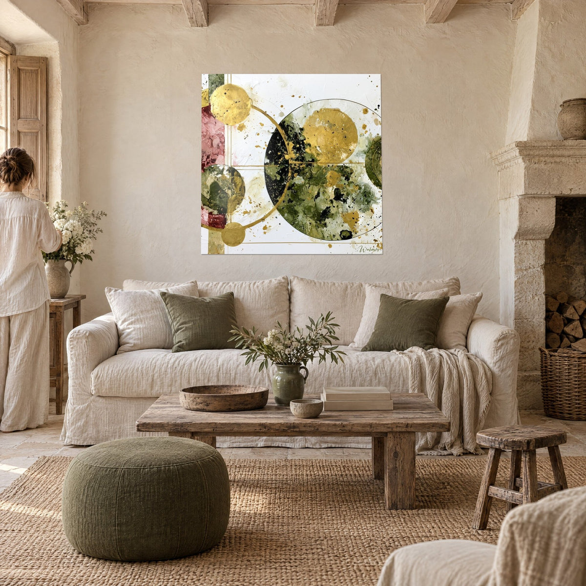 Wall art with golden circles splashed with olive and pink, on a white background, seen from behind by a woman, above a sofa in a bright living room