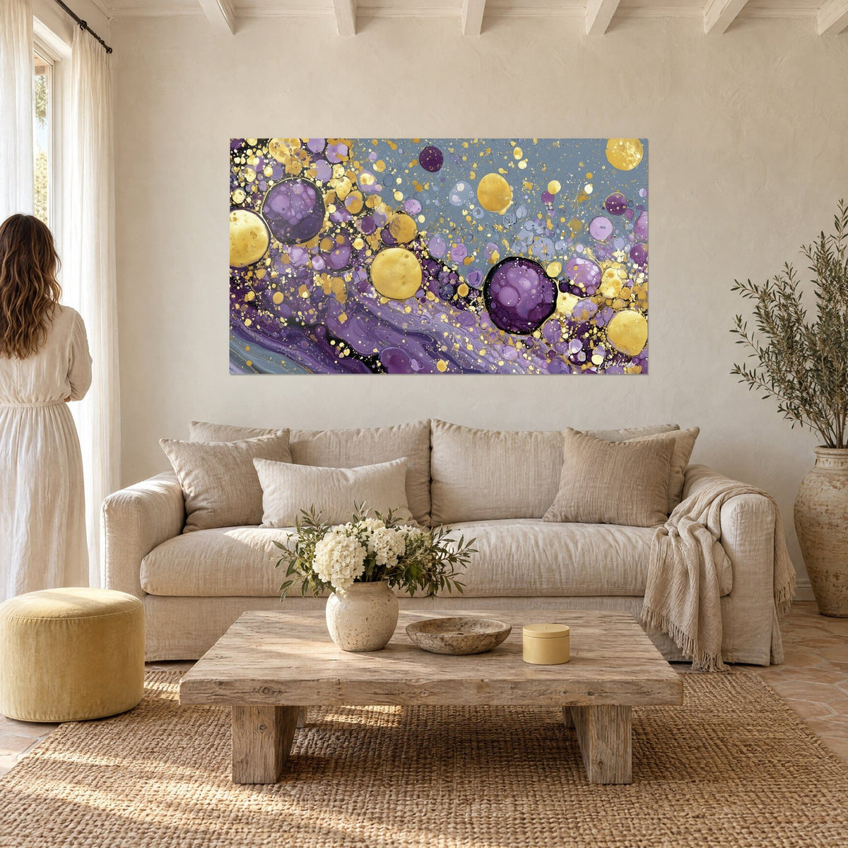 Gemälde mit violetten und goldenen Blasen auf graublauem Grund, eine Frau von hinten betrachtet es über einem Sofa in einem hellen Wohnzimmer