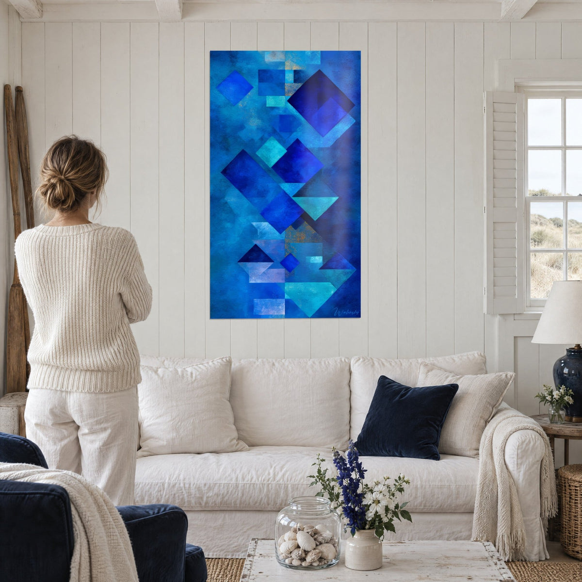 Schilderij ruiten in kobaltblauw en turquoise, vrouw van achteren in een crèmekleurige trui, boven een bank in een lichte woonkamer