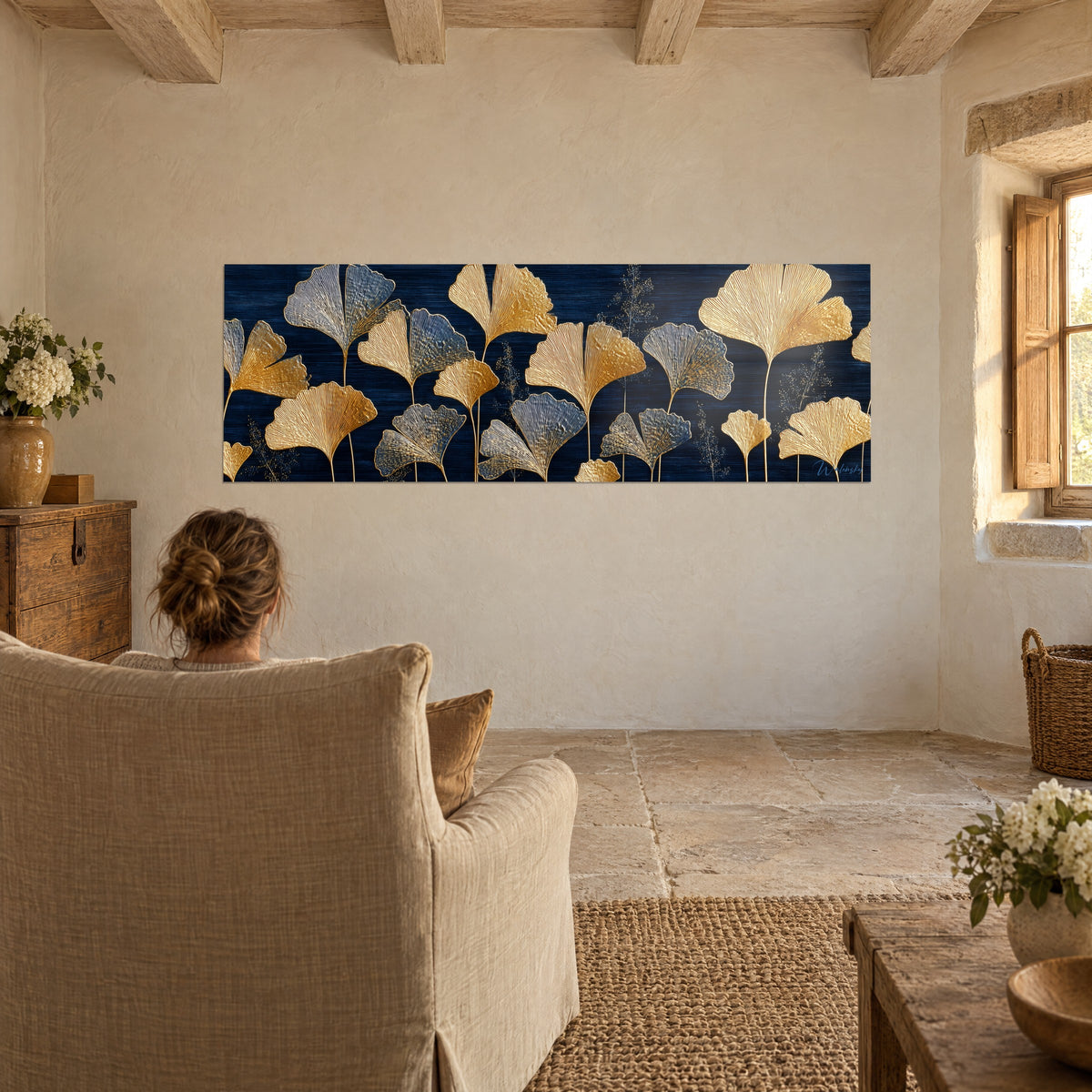 Quadro con foglie di ginkgo nervate in oro, grigio argento e blu navy, ammirato di spalle da una donna su una poltrona del soggiorno