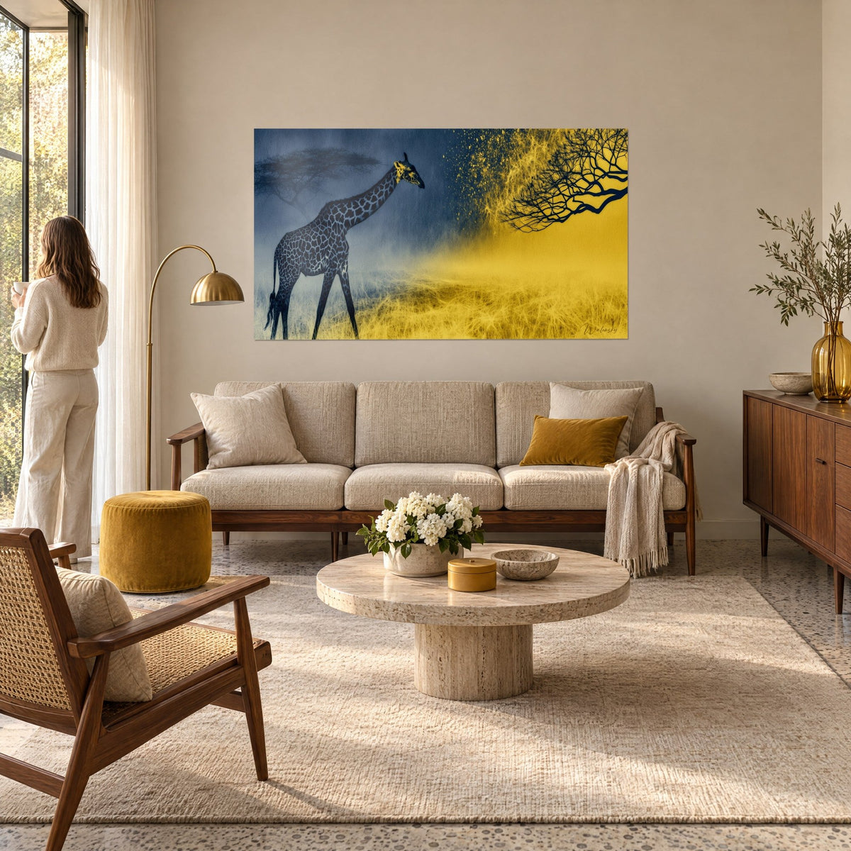 Wall art slender giraffe with navy blue spots, black acacia and yellow savanna grass, woman seen from behind, above a sofa in a bright living room