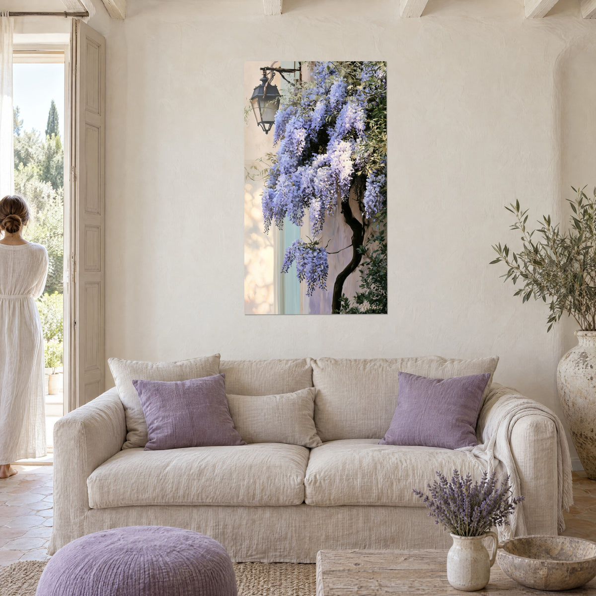 Schilderij met paarse blauweregen trossen en een zwarte lantaarn op een crèmekleurige muur, vrouw van achteren, boven een bank in een lichte woonkamer