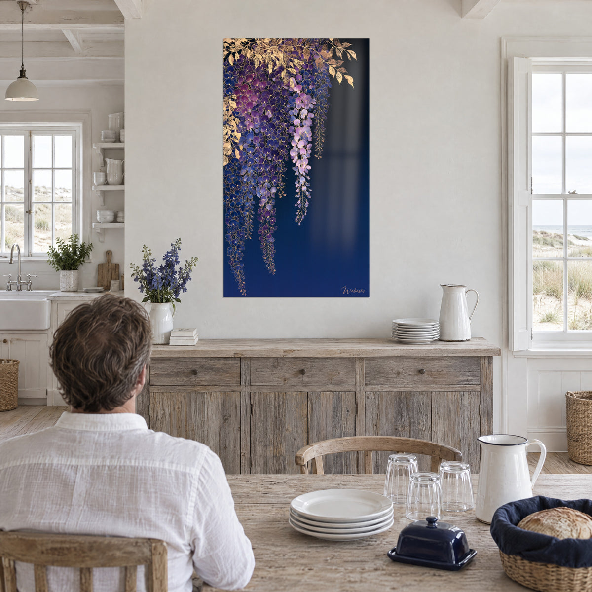 Wall art pale pink and violet floral garland highlighted with gold metallic leaves, a man seen from behind seated at a table in a seaside kitchen