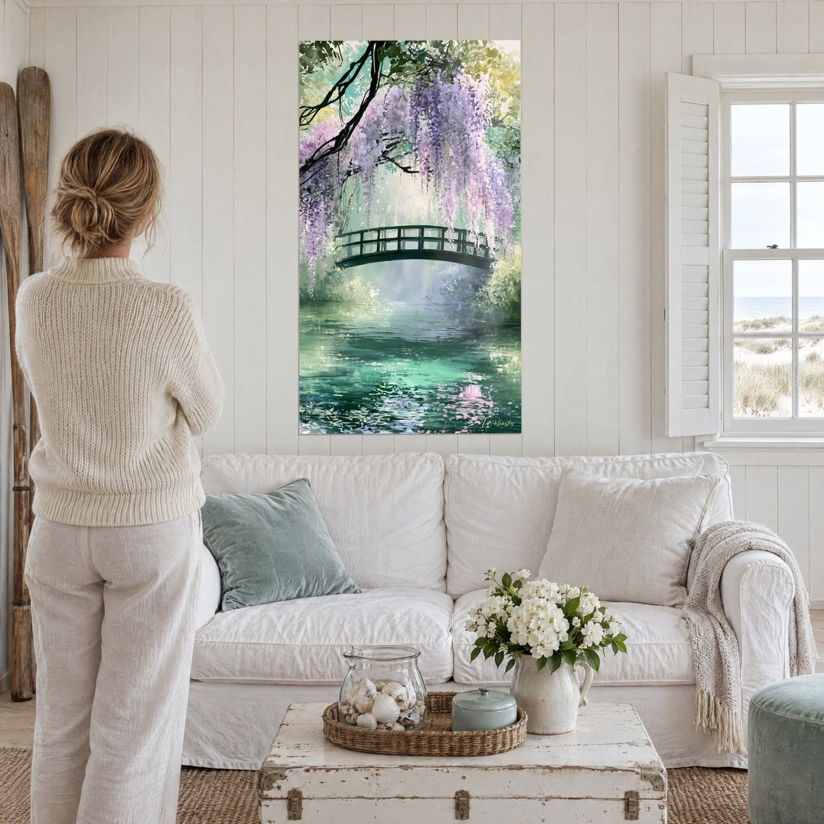Wall art cascading purple wisteria over a black footbridge, emerald green water, woman seen from behind above a sofa in a bright living room