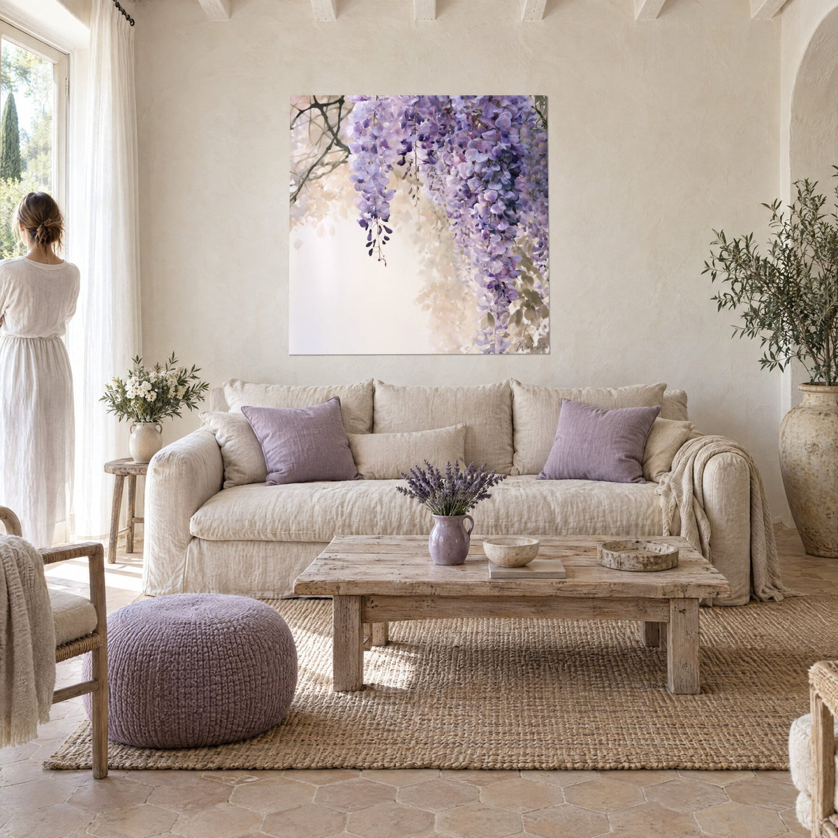 Quadro glicine in grappoli lilla e viola su fondo avorio, una donna in abito bianco di spalle, sopra un divano in un soggiorno luminoso