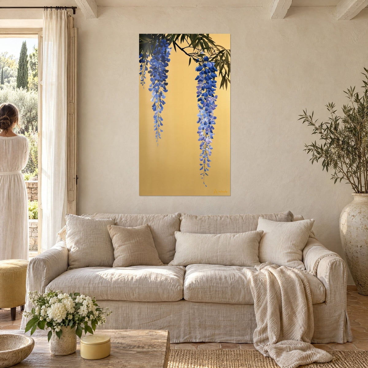 Wall art with cobalt blue wisteria clusters on a gold background, a woman seen from behind admires the canvas above a sofa in a bright living room