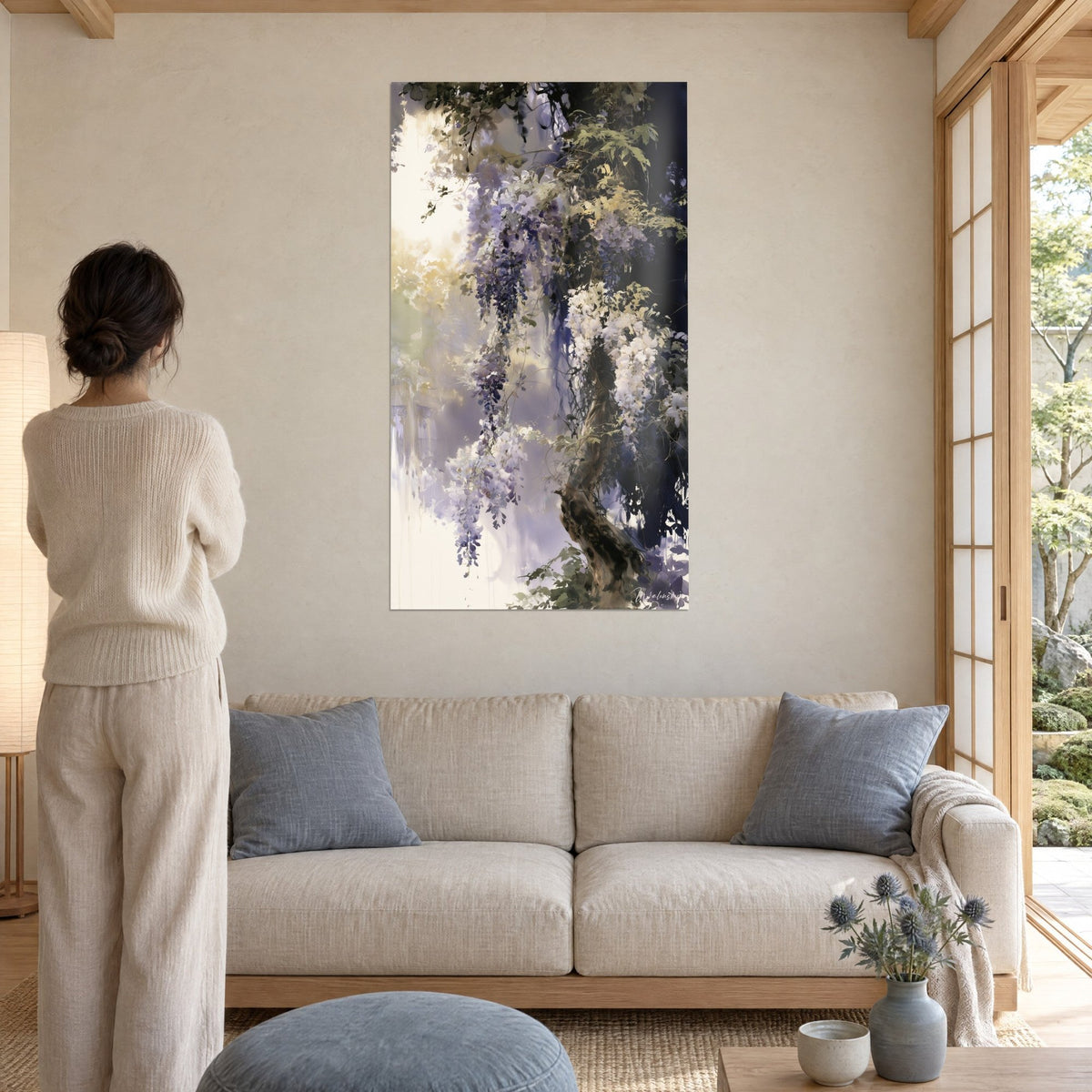 Quadro glicine ad acquerello, grappoli malva e bianchi su tronco bruno, sfondo blu notte, donna di schiena sopra un divano in un soggiorno luminoso