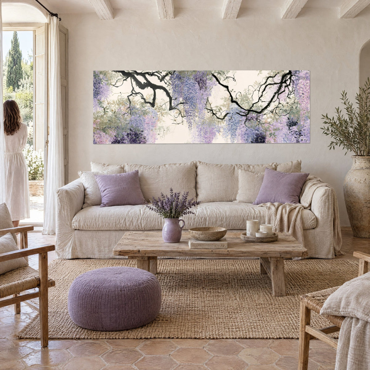 Wall art with cascading clusters of mauve and pink wisteria beneath black branches, woman seen from behind, above a sofa in a bright living room