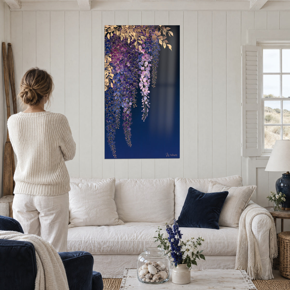 Wall art wisteria in violet and gold clusters on a deep midnight blue background, woman seen from behind, above a sofa in a bright living room