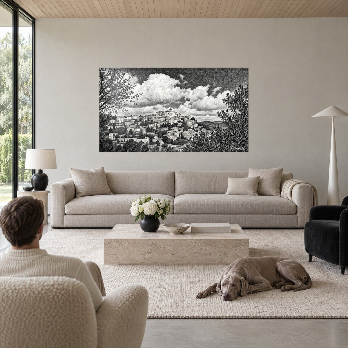 Wall art hilltop village in black and white stippling, puffy white clouds, above a sofa in a bright living room