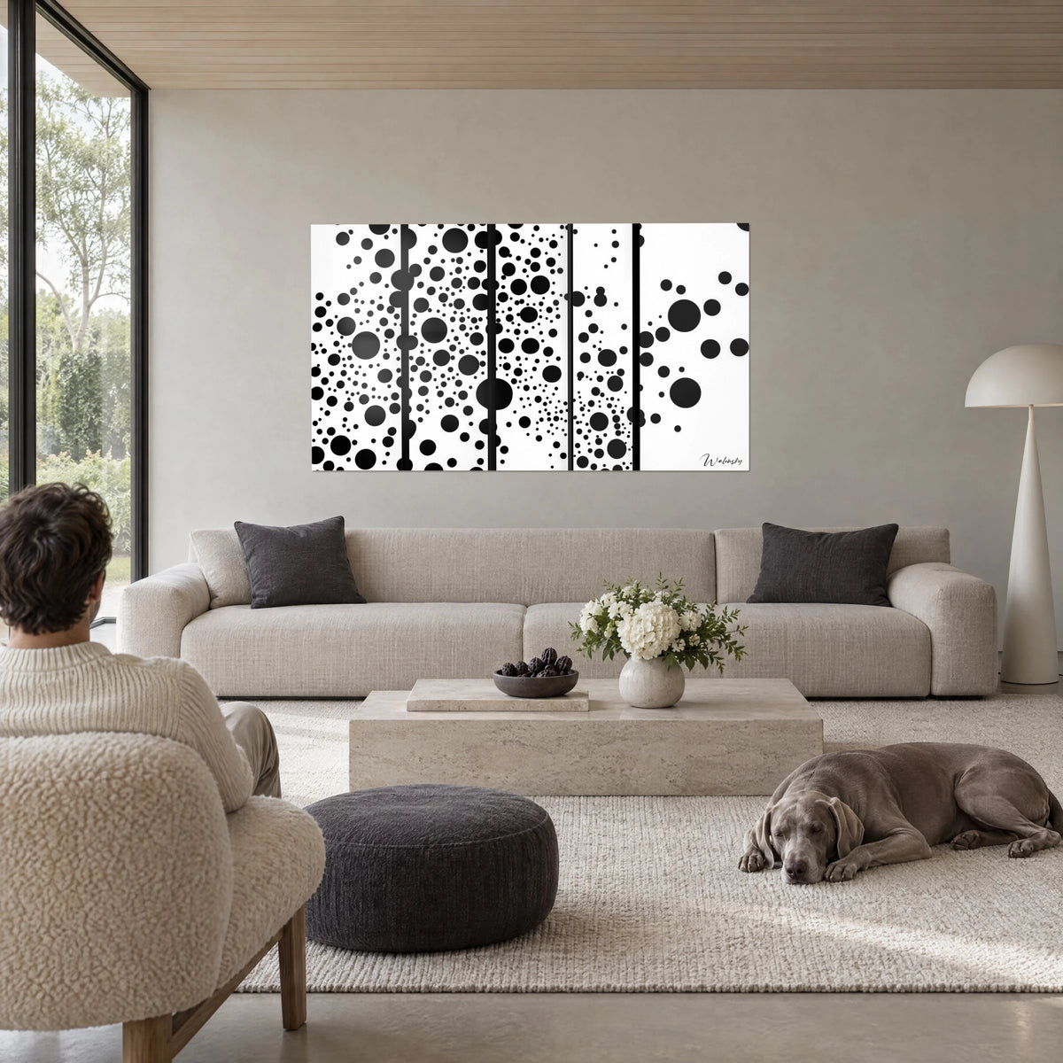 Tableau pois noirs de tailles variées sur fond blanc, cinq panneaux, personne de dos et chien gris au-dessus d'un canapé dans un salon lumineux