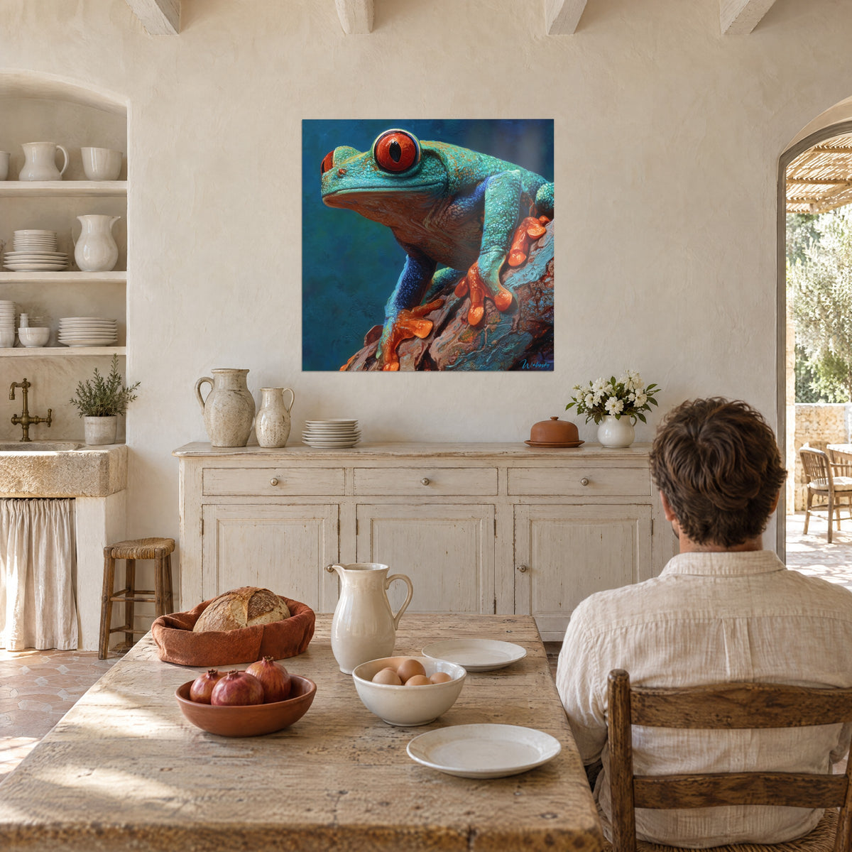 Schilderij turquoise en rode boomkikker op een ruwe tak, man van achteren aan tafel voor een Provençaalse keuken van blonde steen