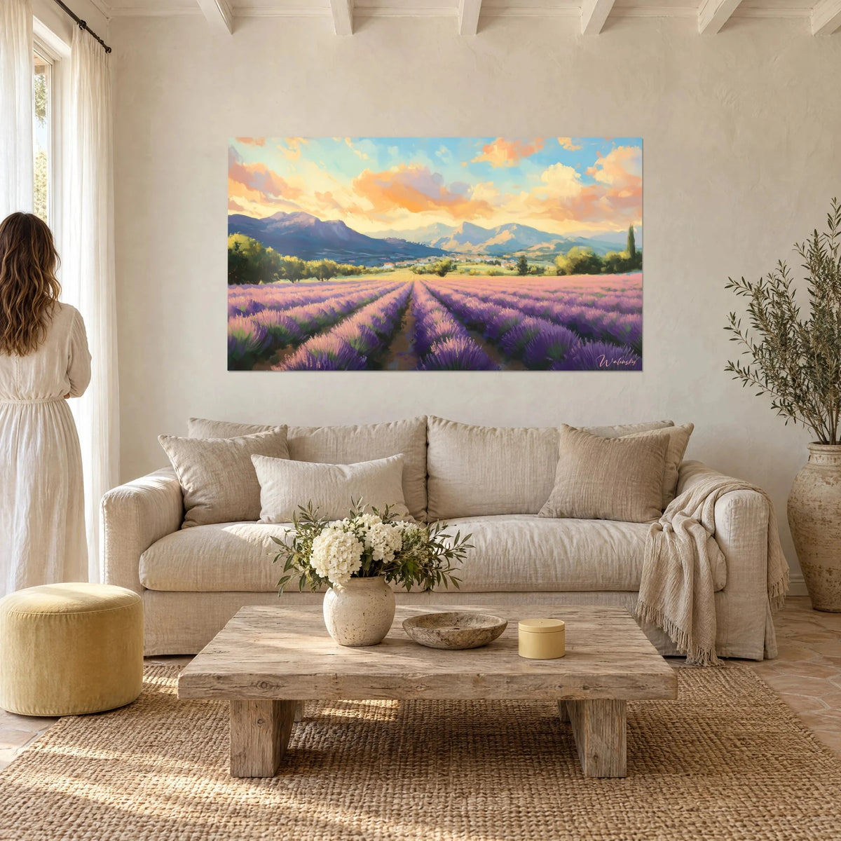 Wall art lavender field in purple rows beneath orange and cream clouds, facing a woman seen from behind, above a sofa in a bright living room