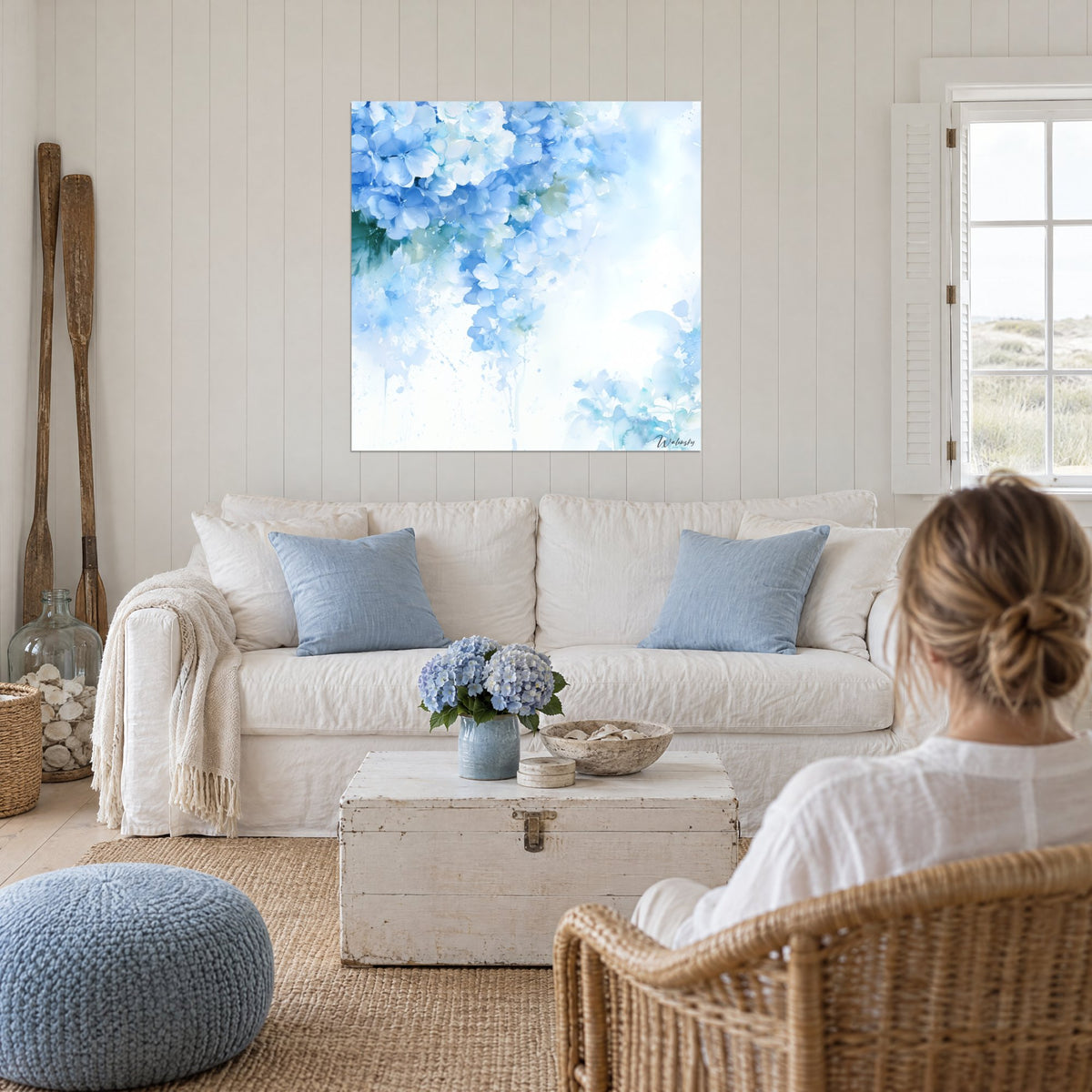 Quadro di ortensie blu e bianche ad acquerello, leggere colature e fondo avorio, sopra un divano in un soggiorno luminoso