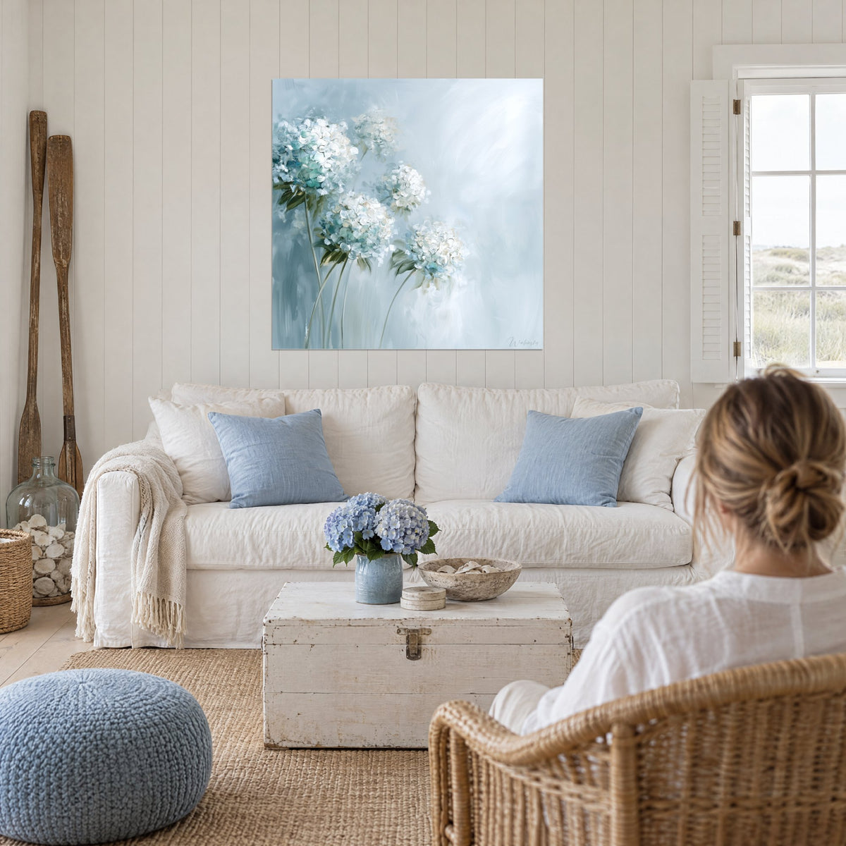 Wall art of white hydrangeas in dense, thick clusters, dark green leaves on powdery blue mist, above a sofa in a bright living room