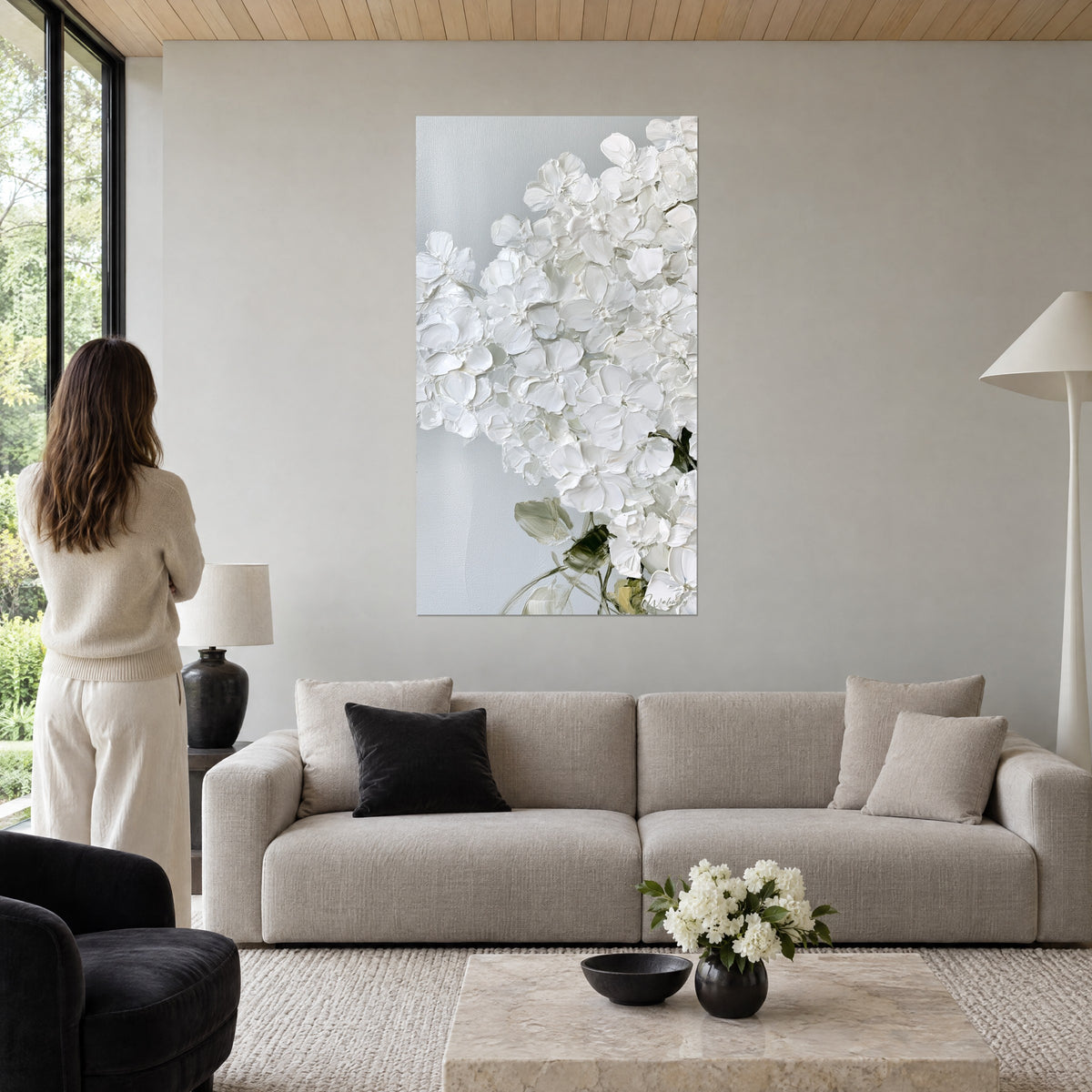 Wall art white hydrangea in relief, ivory petals and sage green leaves, woman seen from behind, above a sofa in a bright living room