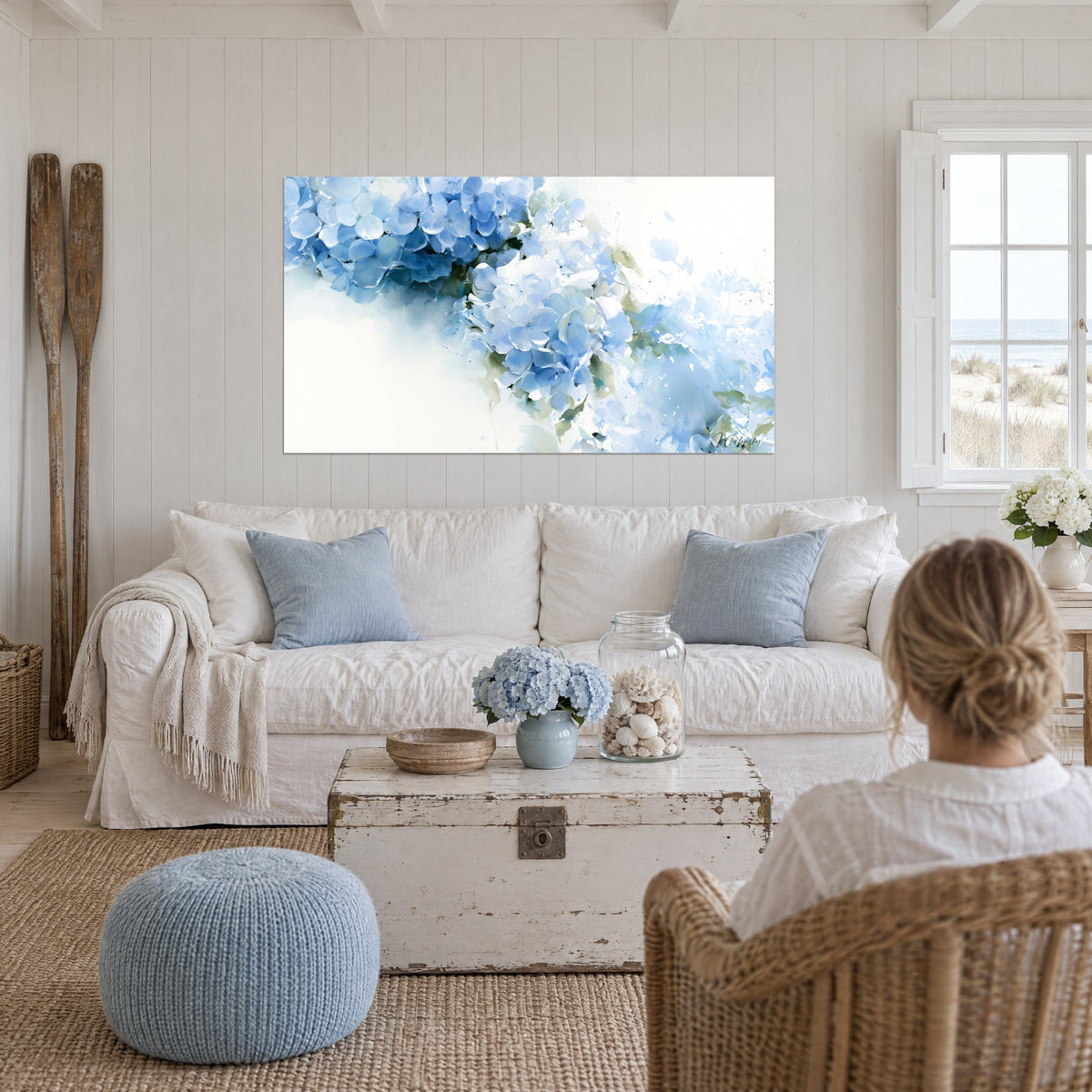 Wall art with sky-blue hydrangeas on a diagonal against a white background, airy petals and sage-green leaves, above a sofa in a bright living room