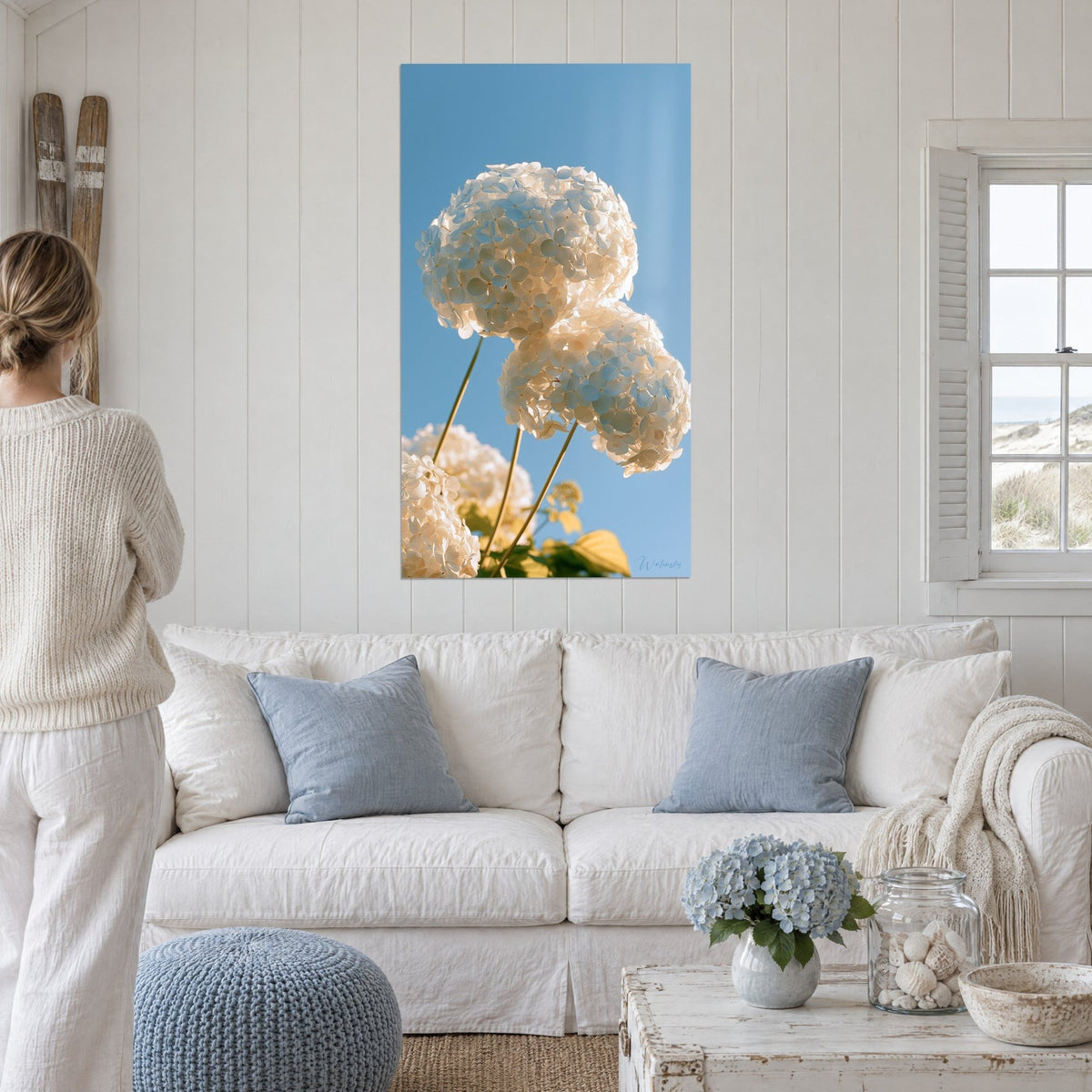 Wall art of white hydrangea balls under a blue sky, golden stems, above a sofa in a bright living room