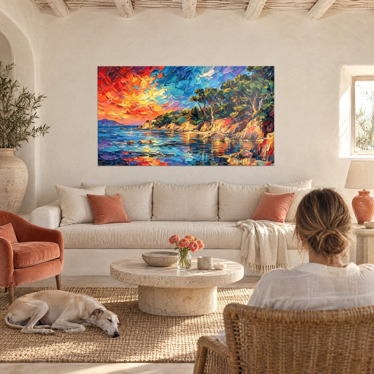 Cuadro de una cala provenzal con cielo naranja y azul, rocas ocres y pinos, dos personas de espaldas y un galgo, encima de un sofá en un salón luminoso