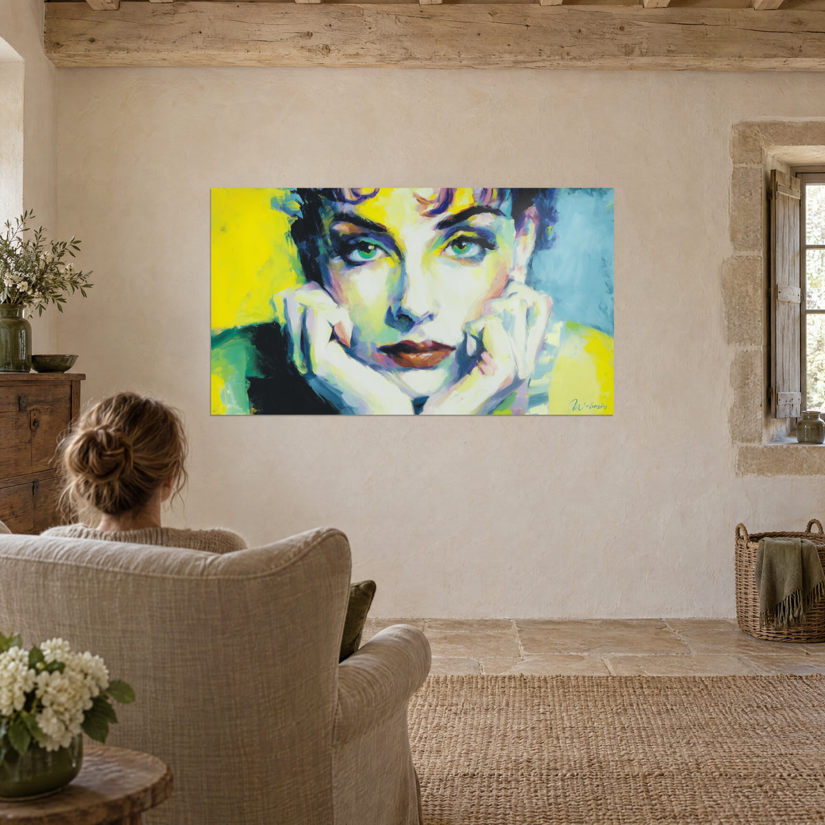 Cuadro figura femenina de mirada verde agua, pinceladas amarillas, azules y violetas, sobre una pared clara junto a un sillón de lino