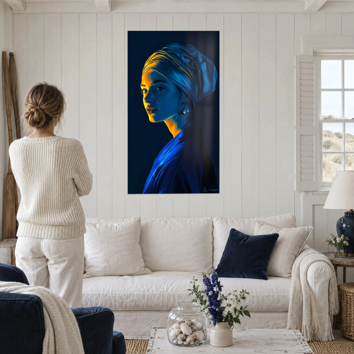 Quadro giovane donna con turbante blu e ocra, orecchino di perla, sfondo notturno, con una donna di spalle sopra un divano in un soggiorno luminoso